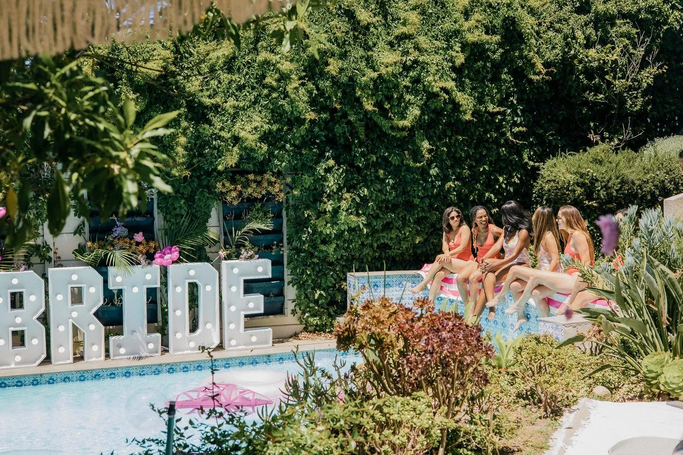 Friends gathered poolside by glowing BRIDE marquee letters at a villa weekend in a lush Cape Town garden